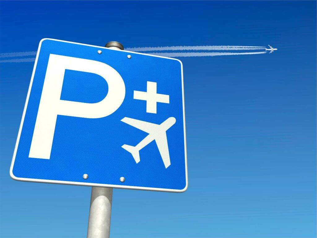 Ein blaues Park-and-Fly-Schild vor klarem, blauem Himmel, während ein Flugzeug am Himmel vorbeifliegt und Kondensstreifen hinterlässt