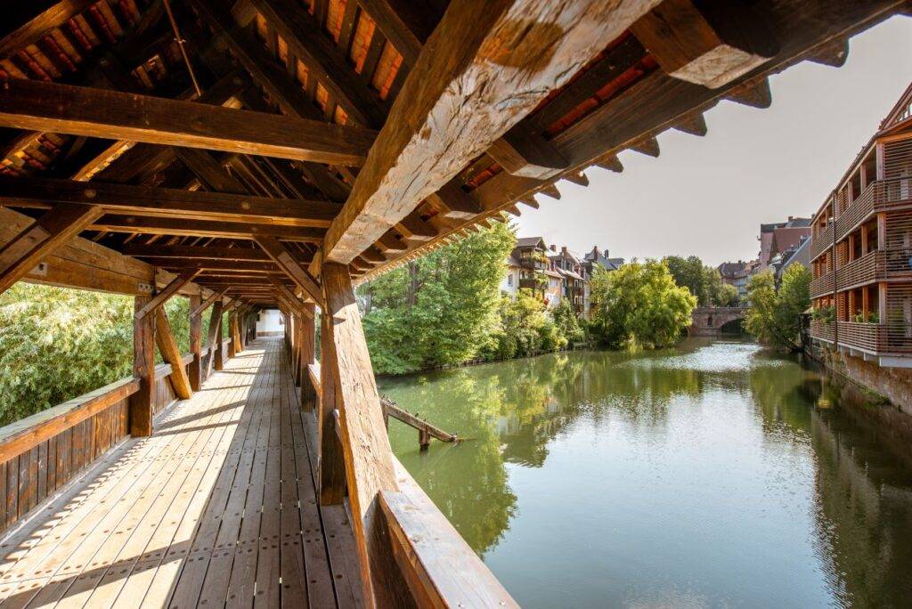 Ein überdachter Holzsteg, der Henkersteg in Nürnberg, führt über den Fluss Pegnitz und bietet einen idyllischen Blick auf das Wasser und die umliegenden Gebäude