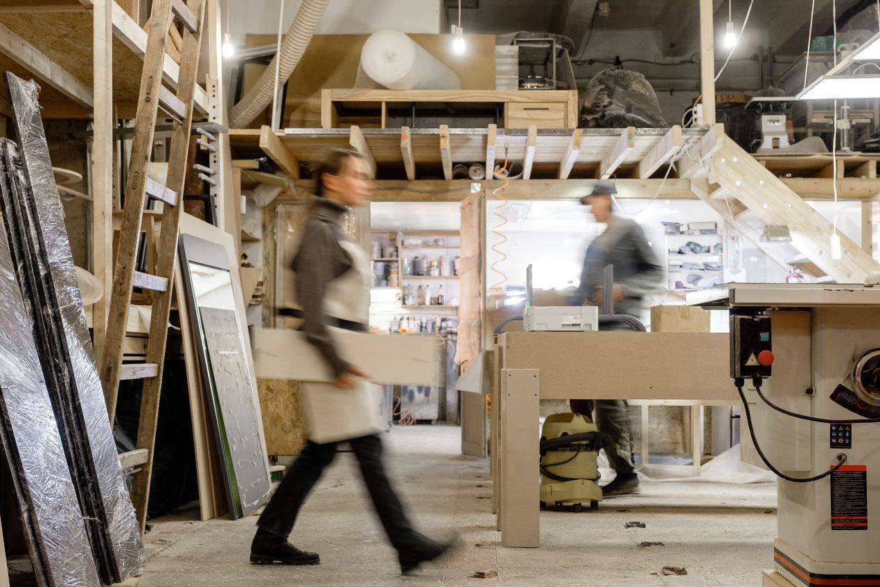 Zwei Handwerker in einer Möbelbau-Werkstatt in Berlin mit Holzmaterialien und Maschinen im Hintergrund – Möbelbau Berlin